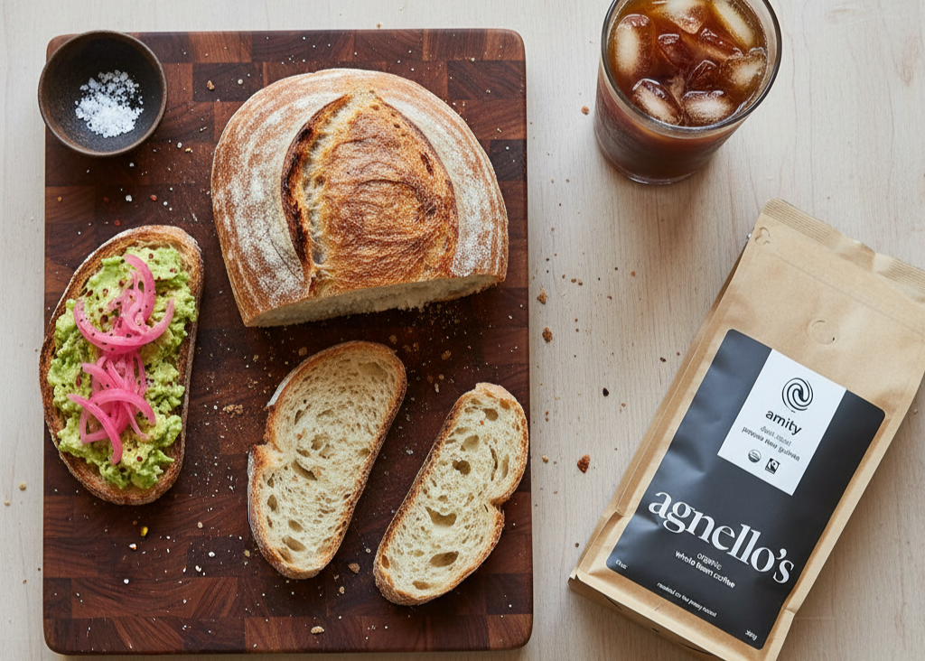 Loaf of bread on a wooden cutting board with a slice cut, next to a glass of iced coffee and a package of 'agnello's' coffee.